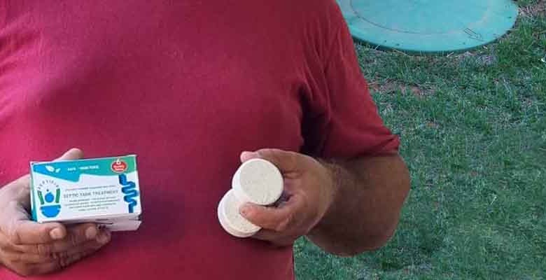 Hands holding a Septifix box and two white septic tablets, with a septic tank lid on the lawn behind